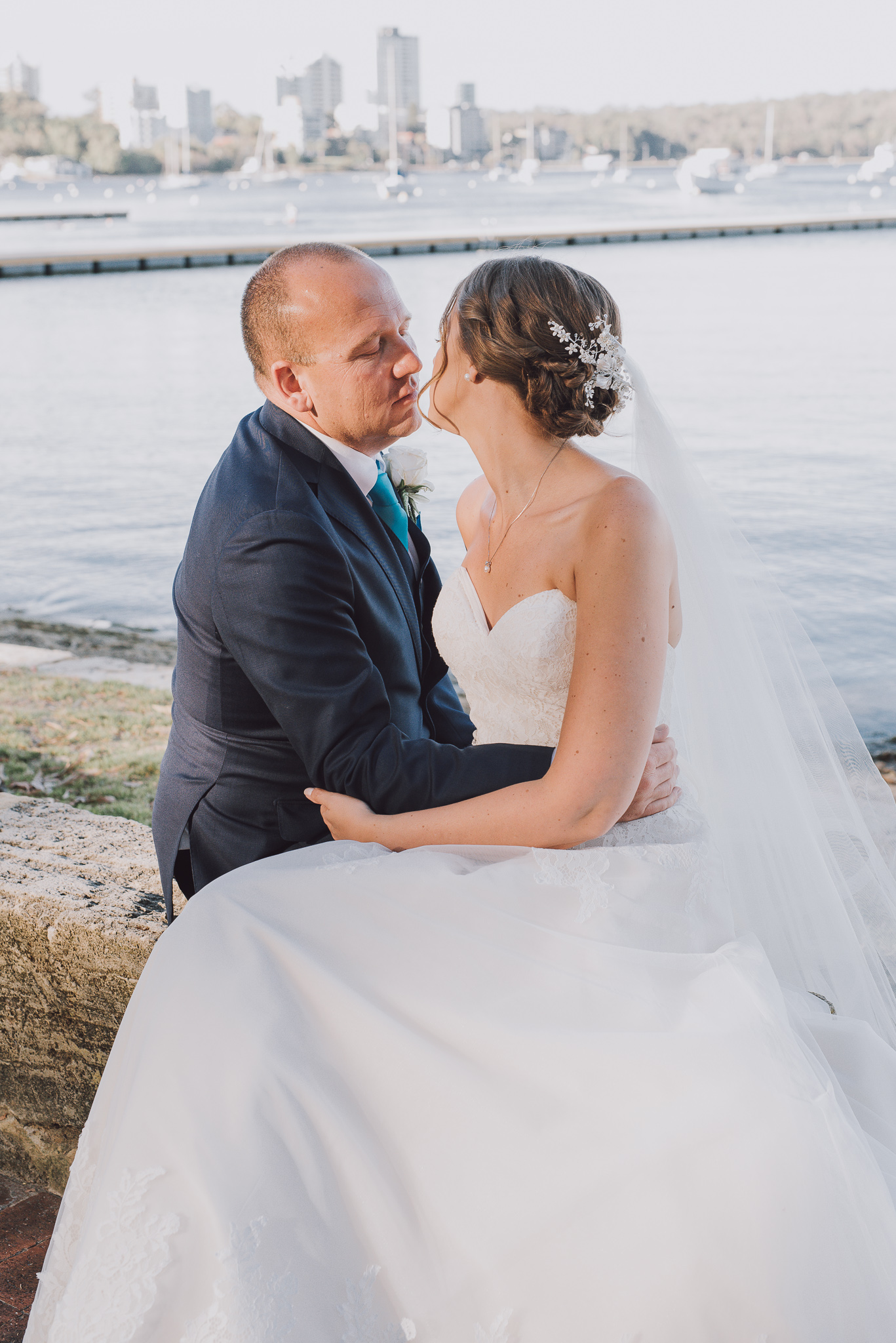 bride and groom about to kiss sitting on a wall next to the river