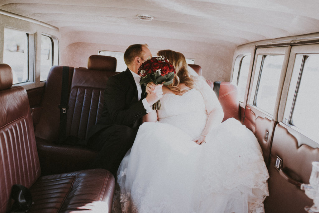 Bride and groom kissing behind their bouquet in wedding car